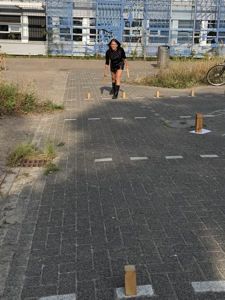 A person is joyfully playing an outdoor game with wooden blocks and sticks on a paved area at Kade West, Marineterrein, Amsterdam